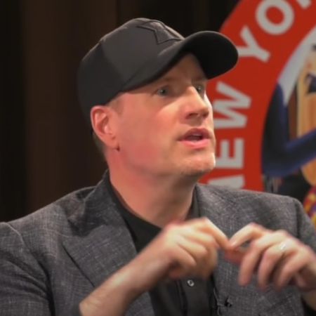 Kevin Feige is sitting on stage wearing a grey suit and a black cap.
